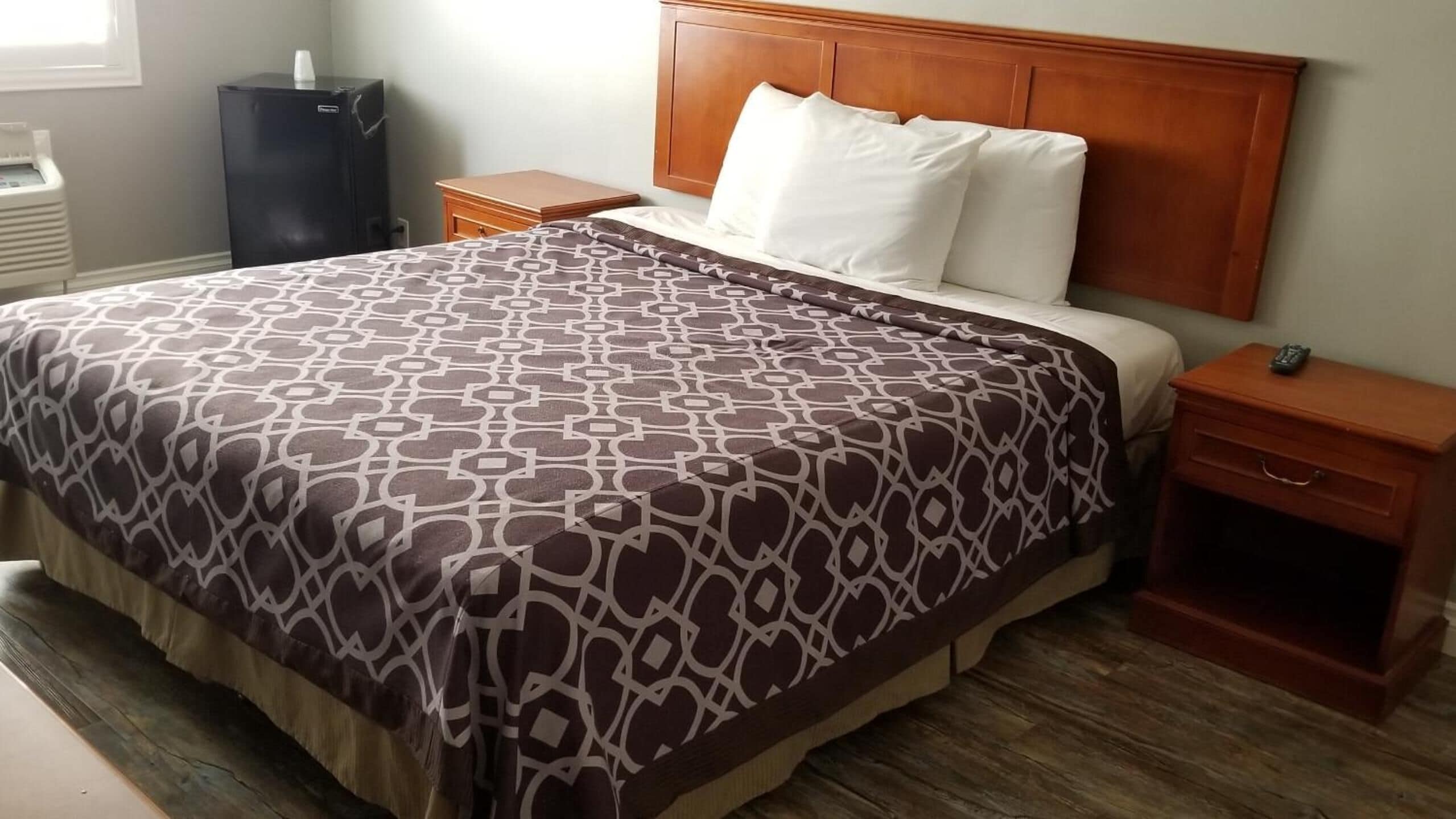 Habitación de hotel con una cama grande, cabecera de madera, almohadas blancas, colcha estampada, mesita de noche con control remoto y un pequeño armario de madera.