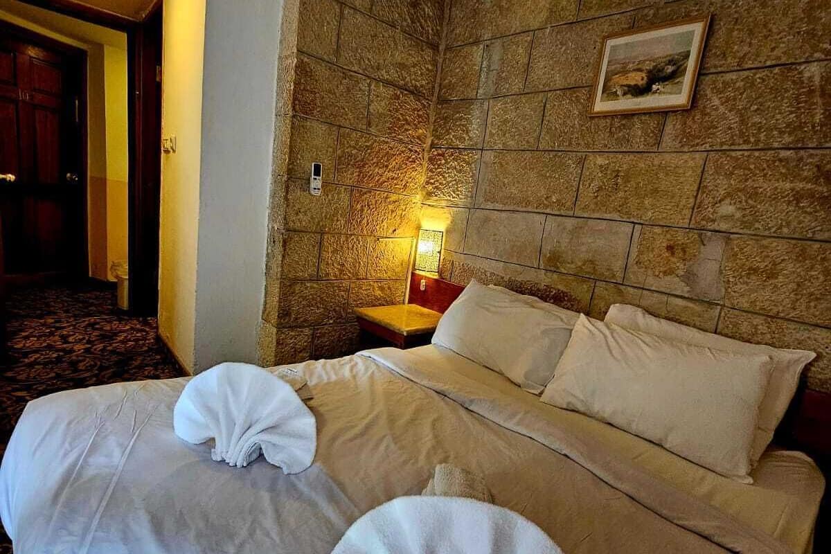 A hotel room with a stone accent wall, a bed with white linens, a nightstand with a lamp, and an air conditioning unit above the bed.