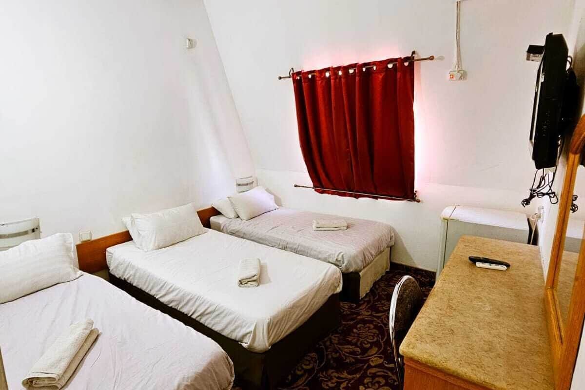 A hotel room with two single beds, a small desk, a telephone, and a window with red curtains.
