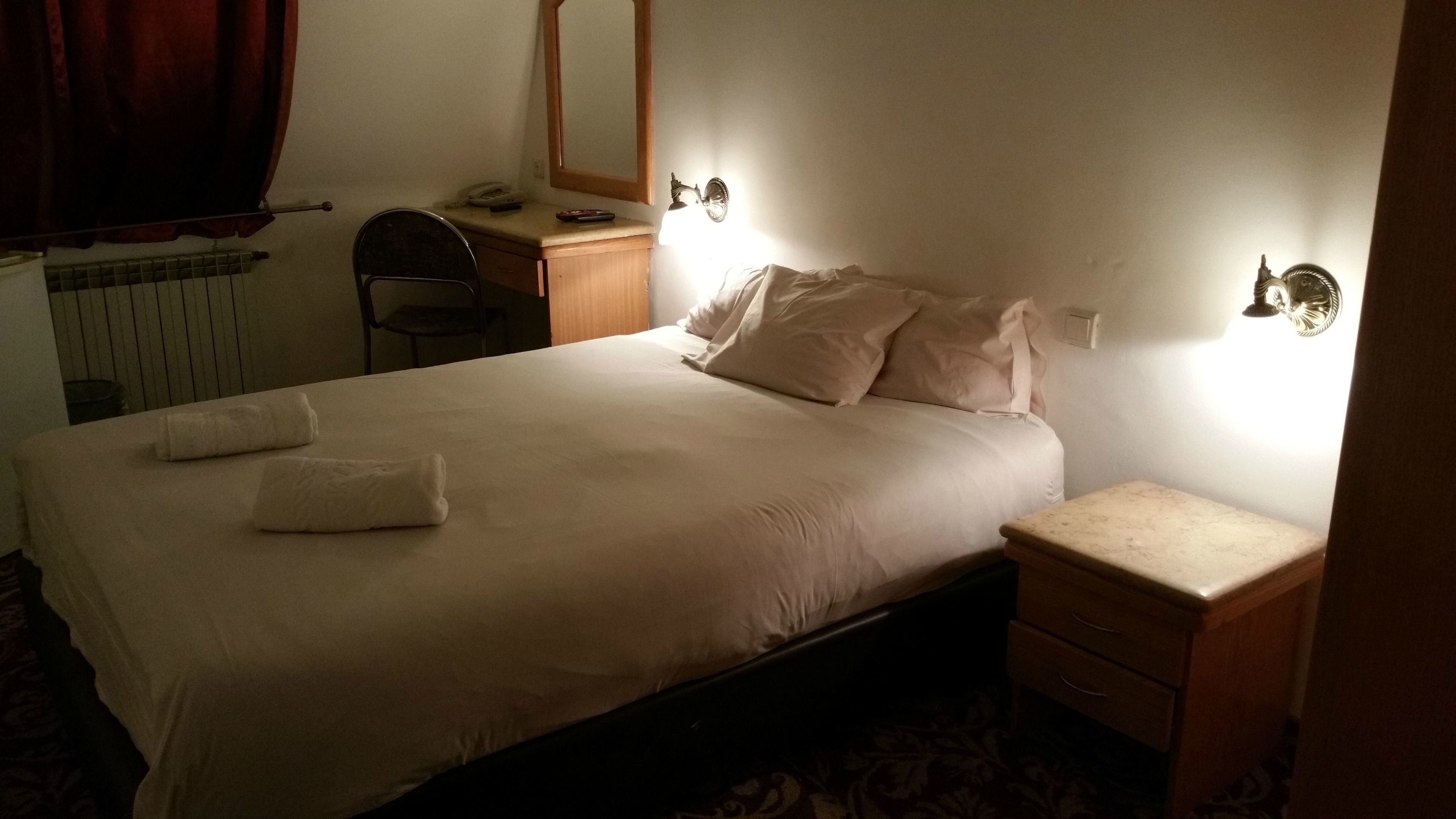 A neatly made bed with two pillows and two folded towels, a bedside table with a lamp, and a chair in a room with a radiator and a mirror on the wall.