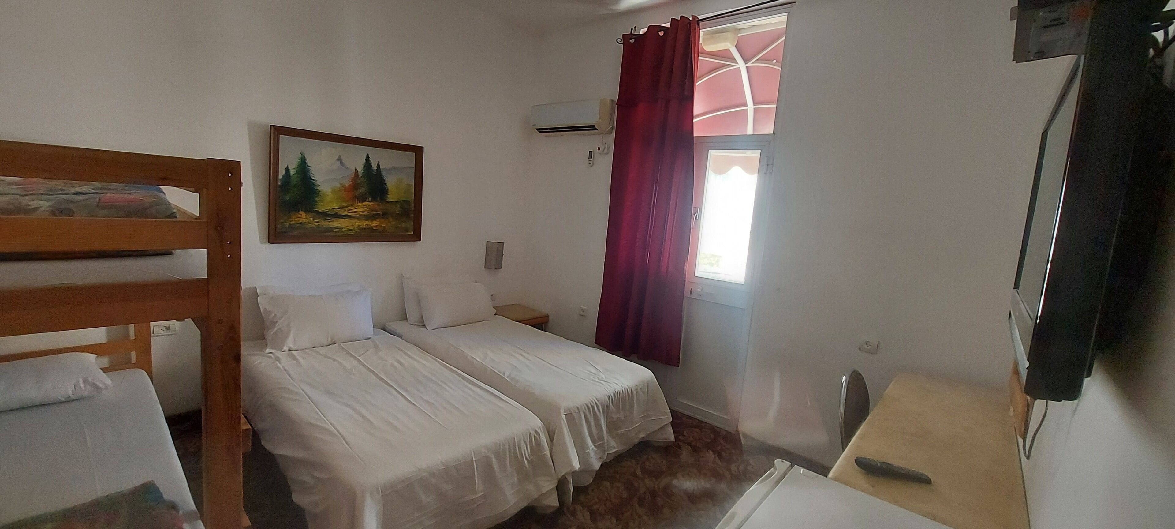 A room with two single beds, a wooden bunk bed, a painting on the wall, a window with red curtains, and a small table with a chair.