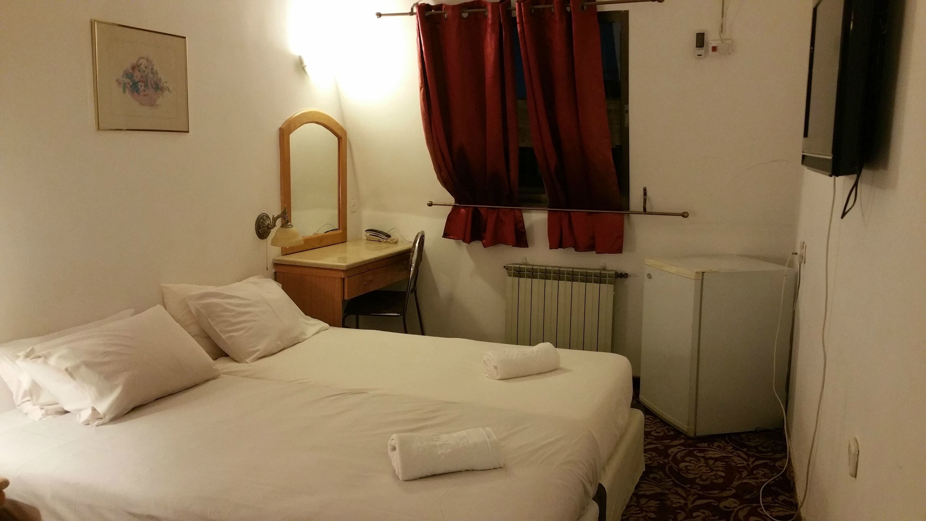 A hotel room with a bed, a desk, a mirror, a radiator, and a television.
