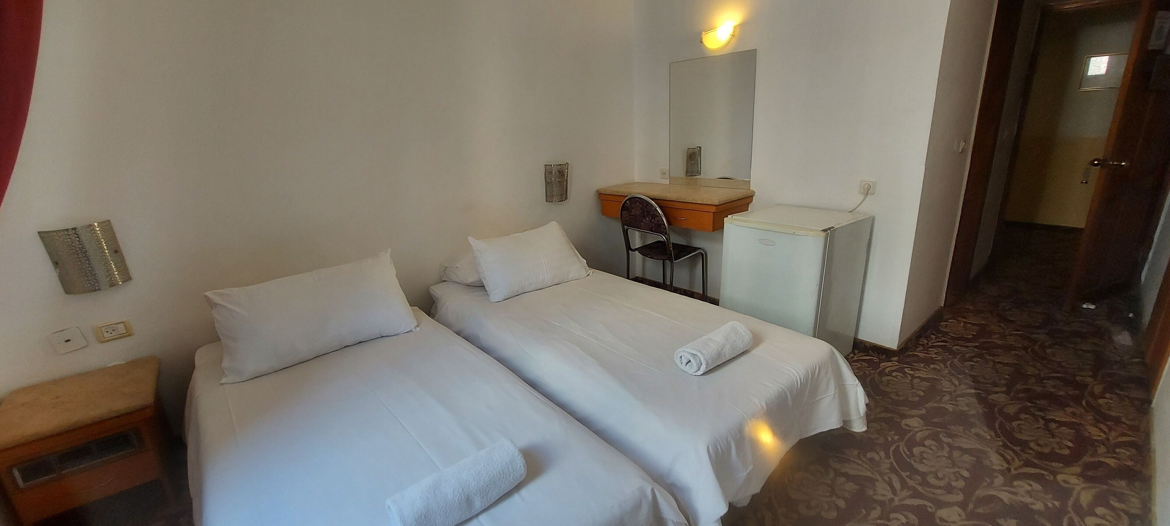 Habitación de hotel con dos camas individuales, un escritorio pequeño, una silla, un espejo y suelo alfombrado.