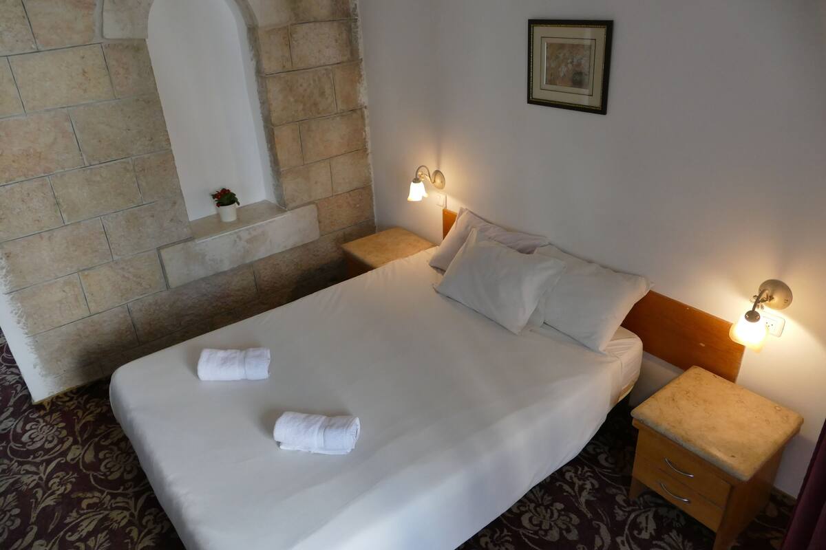 A neatly made bed with white linens and two towels, a wooden nightstand with a lamp, and a stone wall with a white archway.
