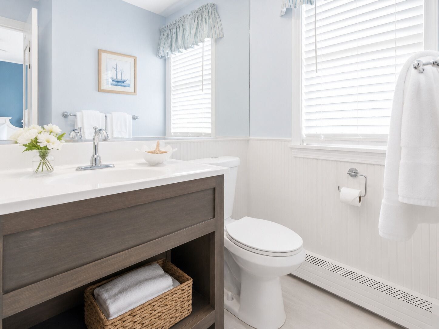 A bathroom with a white sink, mirror, and toilet. There is a framed picture on the wall and a window with blinds.