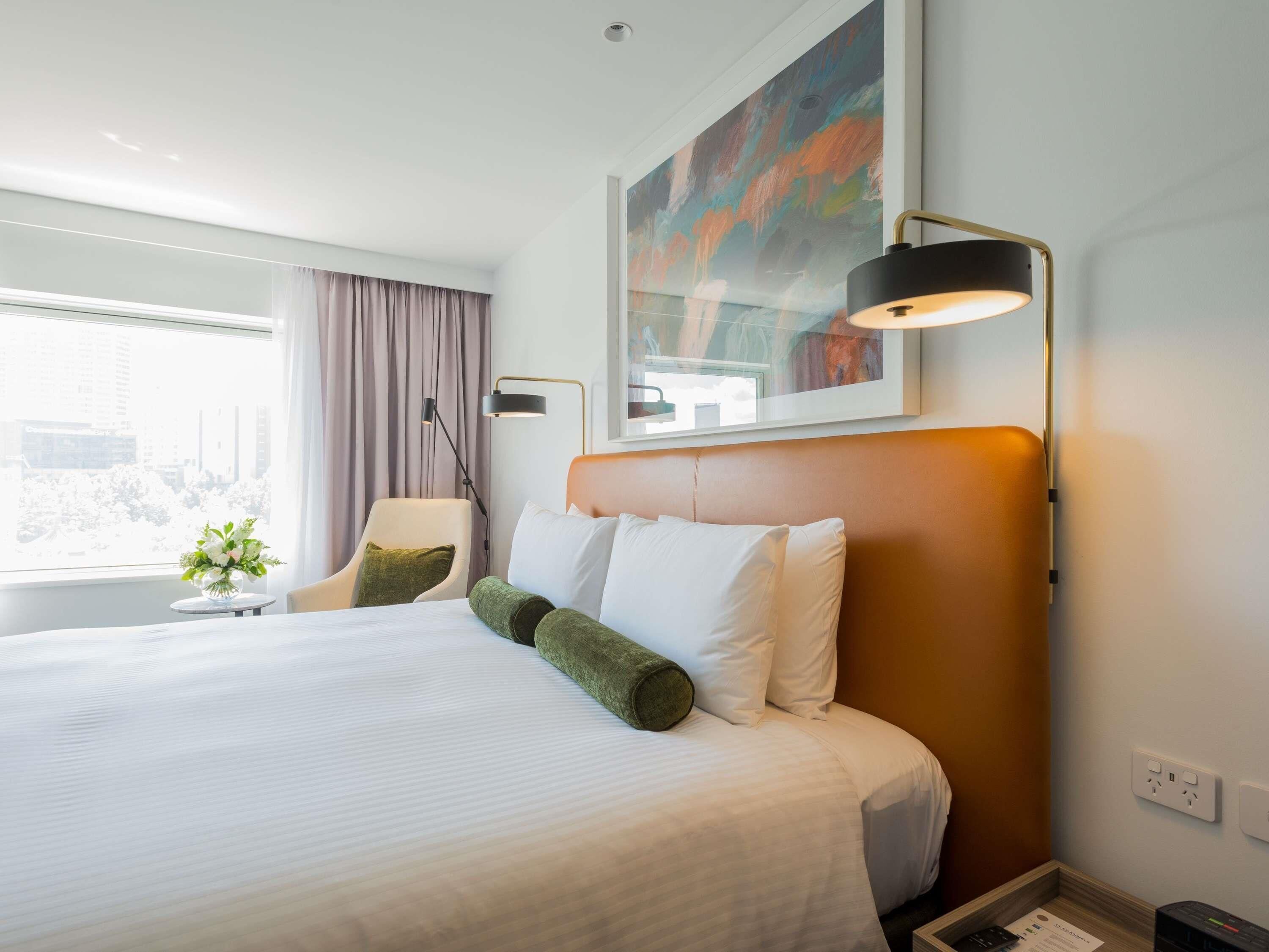 Habitación de hotel con una cama grande, un escritorio, una silla, una ventana con cortinas, un cuadro en la pared y una lámpara.