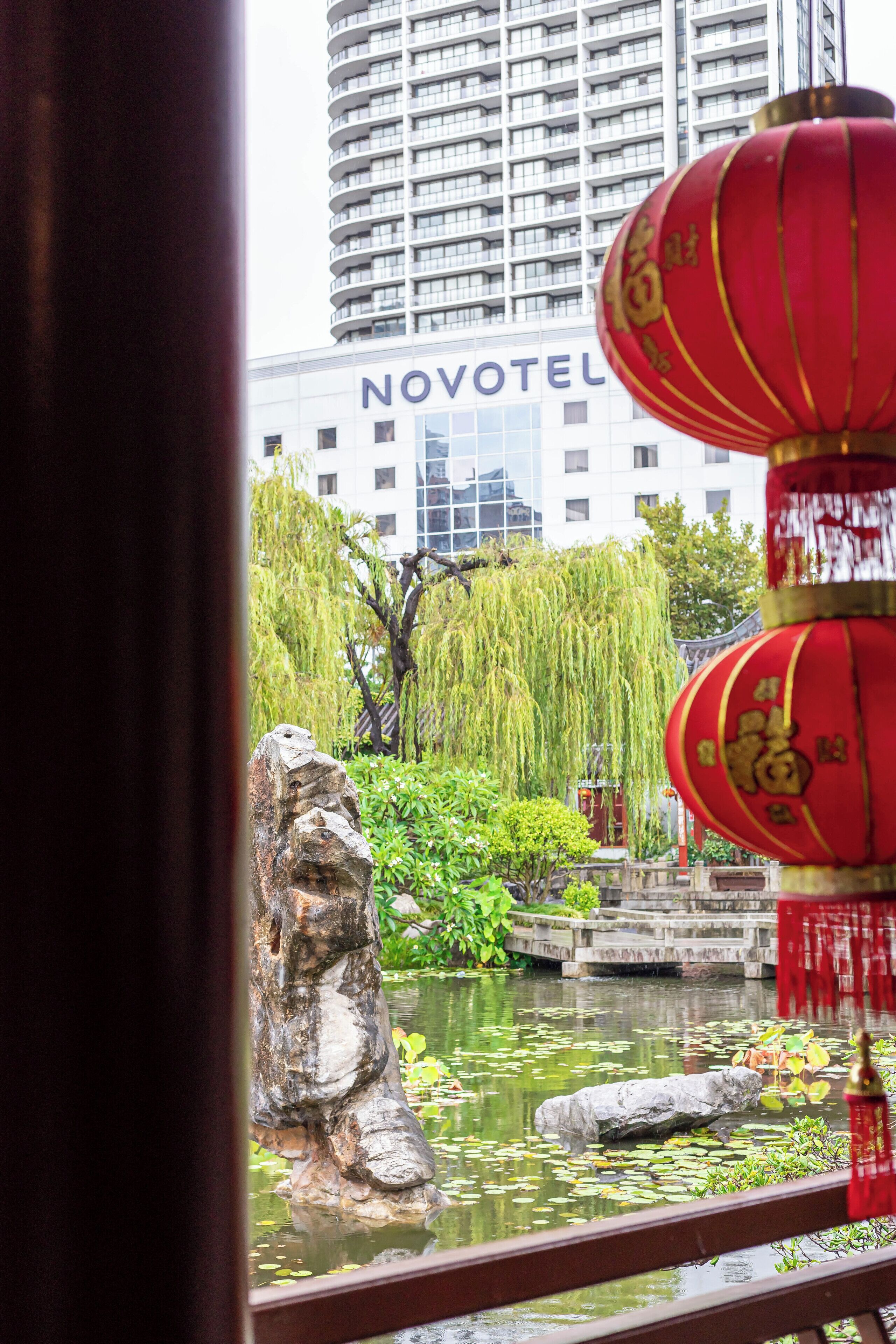 A view of a pond with a rock formation, surrounded by greenery, with a Novotel hotel in the background.