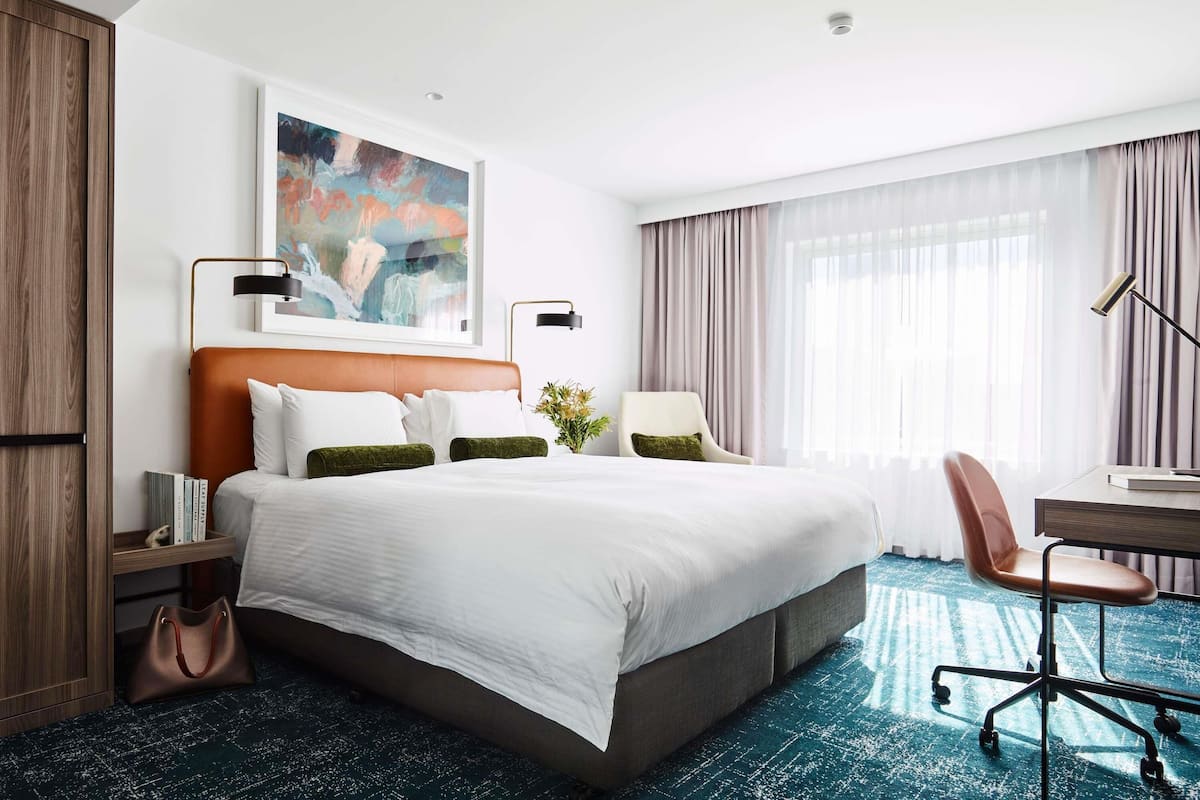 A hotel room with a large bed, a desk, a chair, a wardrobe, and a painting on the wall.