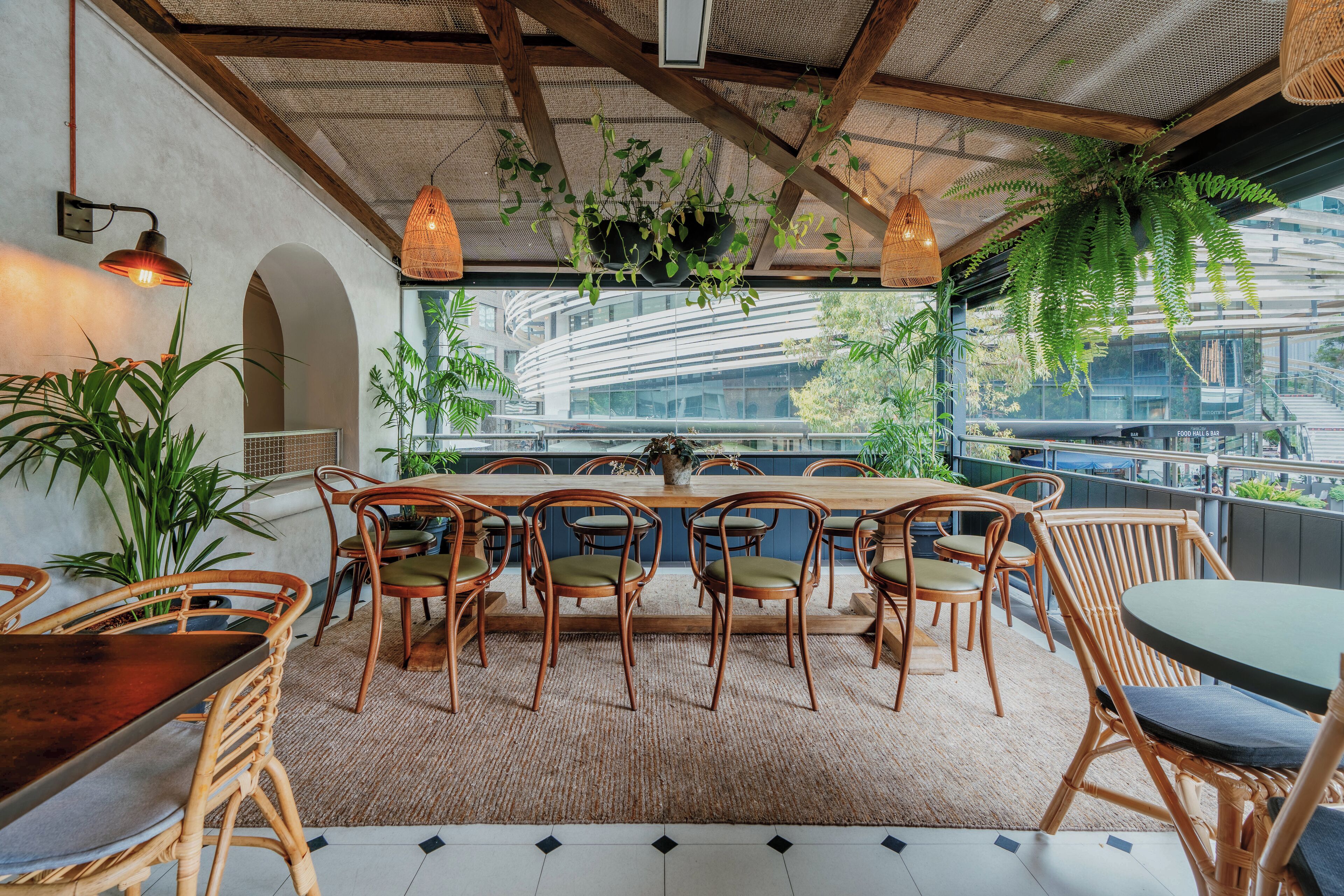 A dining area with a long wooden table and wicker chairs, surrounded by potted plants and hanging greenery.