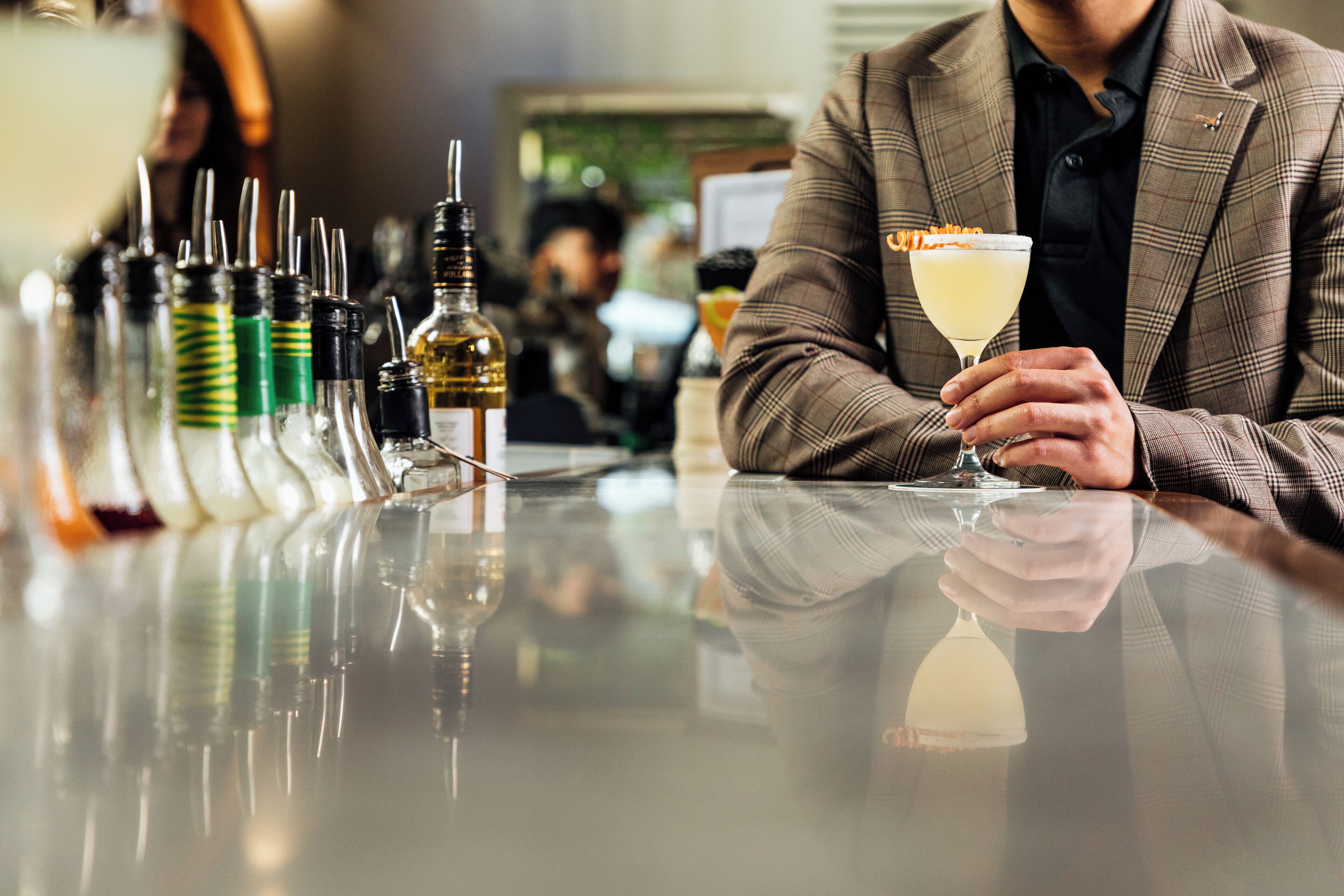 A person seated at a bar with a drink in hand, various bottles and a glass in front.