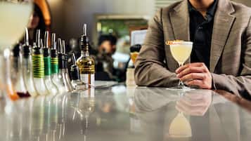 A person seated at a bar with a drink in hand, various bottles and a glass in front.