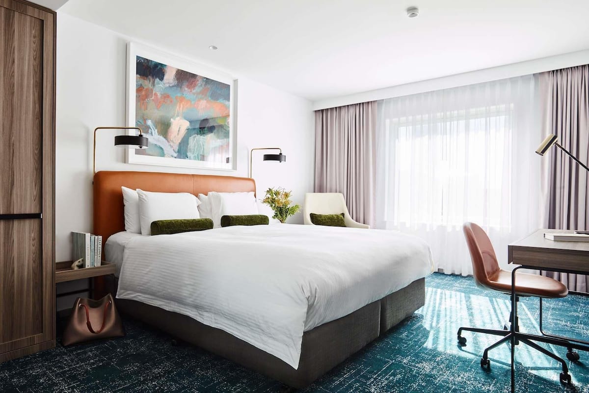 Habitación de hotel con una cama grande, un escritorio, una silla, una lámpara, un cuadro y una ventana con cortinas.