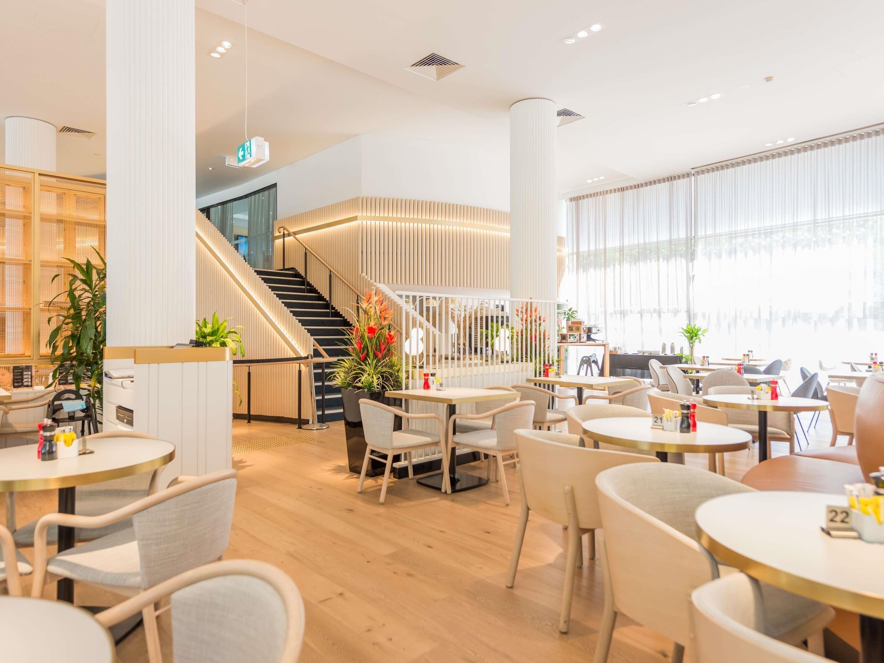 A modern restaurant interior with wooden flooring, round tables, and chairs, a staircase, and large windows.