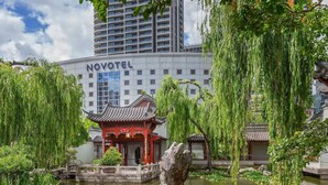 A traditional Chinese pavilion with a pond, surrounded by lush greenery and a modern Novotel hotel in the background.