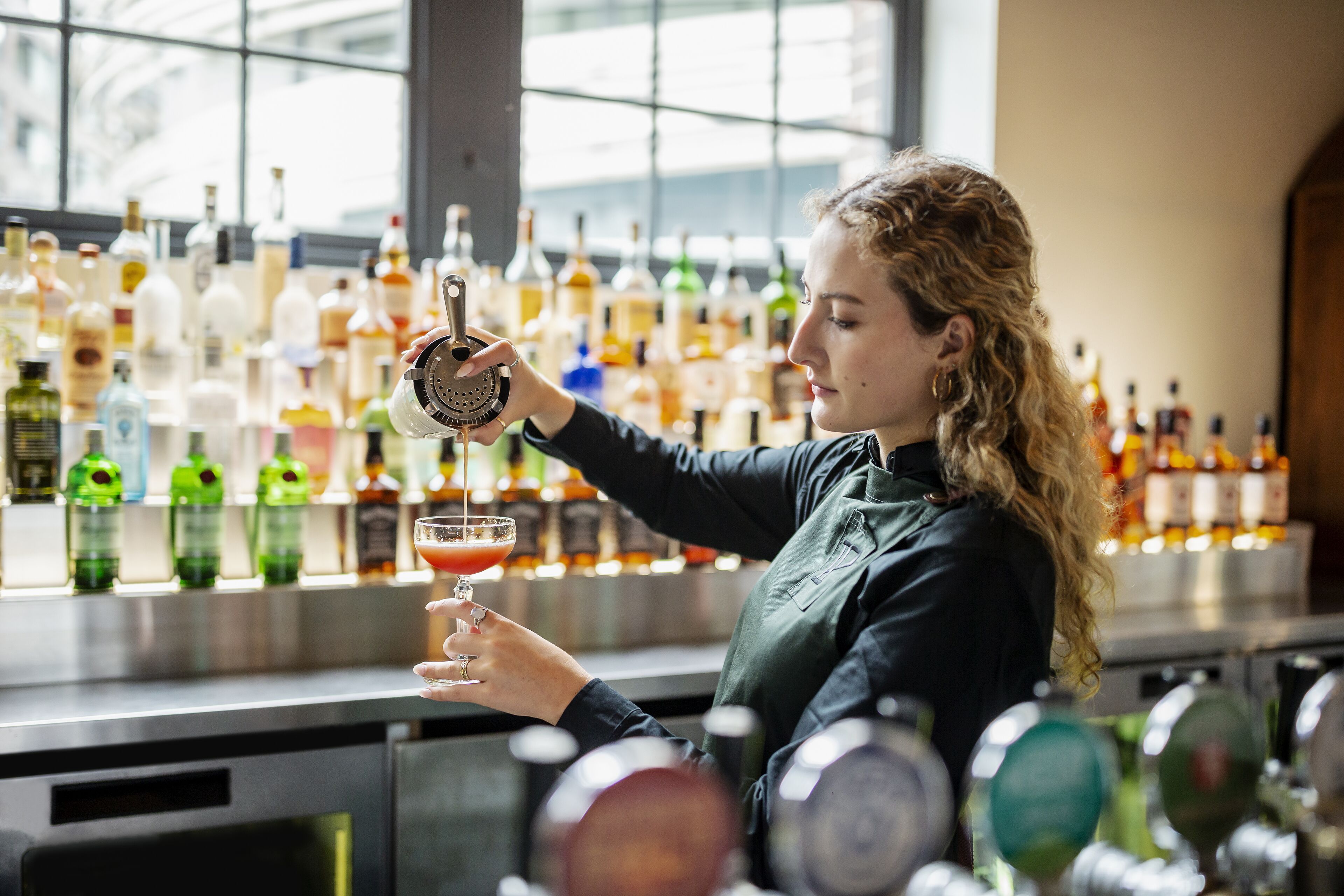 Ein Barkeeper bereitet ein Getränk an der Bar zu, im Hintergrund sind verschiedene Flaschen zu sehen.