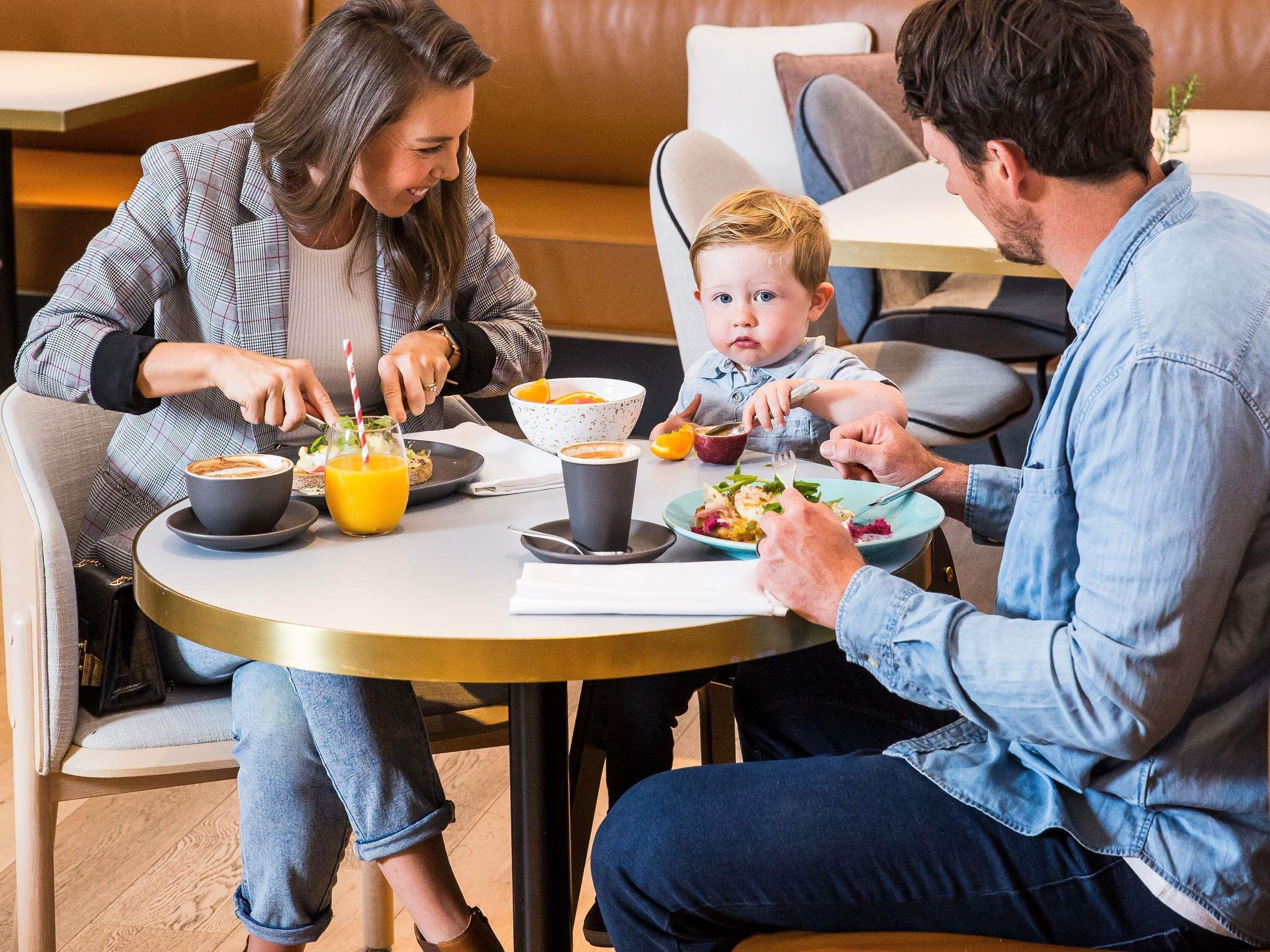 A family dining at a round table with a cup of coffee, a bowl of fruit, and a plate of food.
