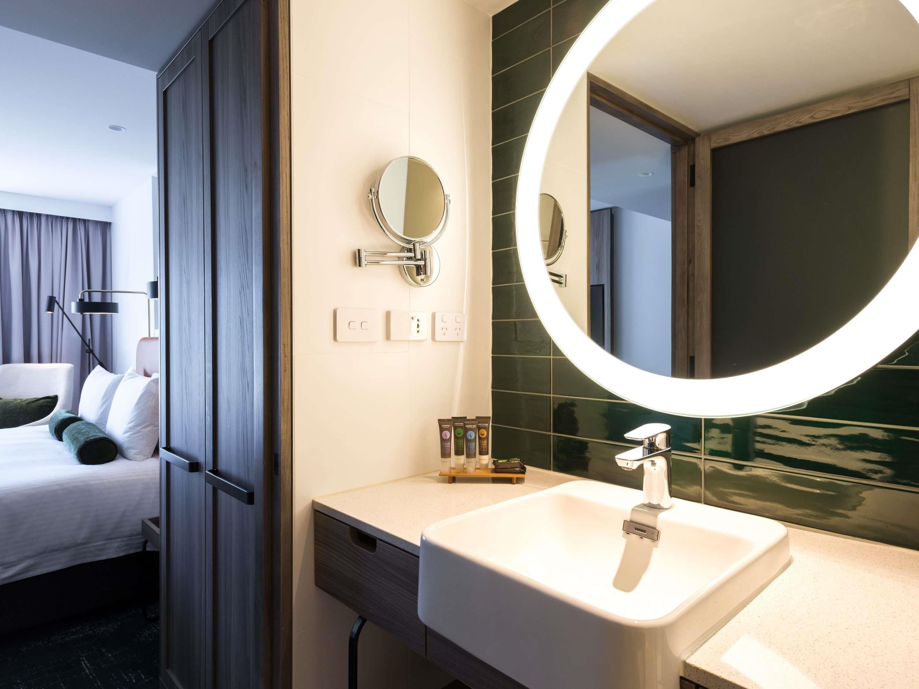 Une chambre d’hôtel avec un lit, une lampe de chevet, un miroir rond, un lavabo et un mur recouvert de carreaux sombres.