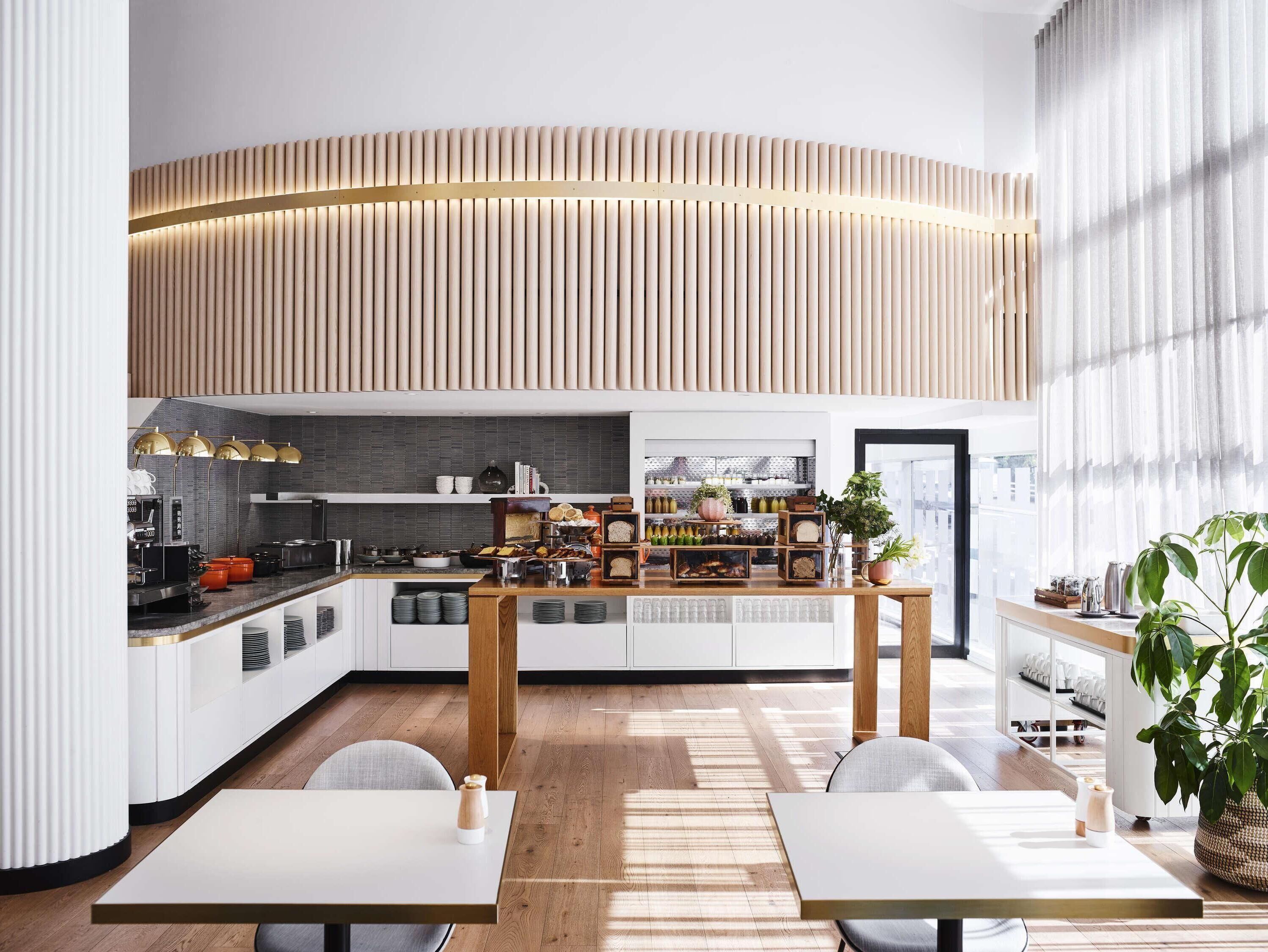 A modern kitchen with a curved wooden panel ceiling, a central island with various food items, and a view of the city through large windows.