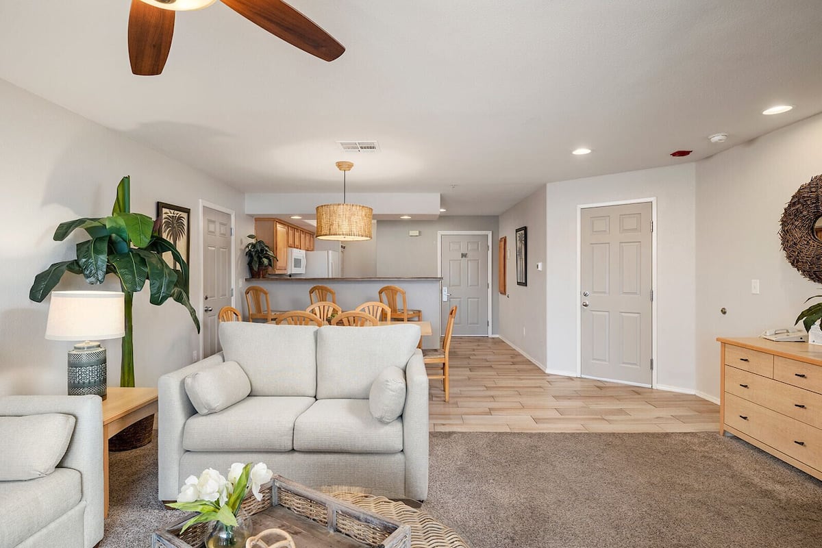 A modern living room with a sofa, a dining table, and chairs. There is a kitchen in the background with a hanging light fixture.