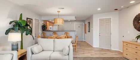 A modern living room with a sofa, a dining table, and chairs. There is a kitchen in the background with a hanging light fixture.