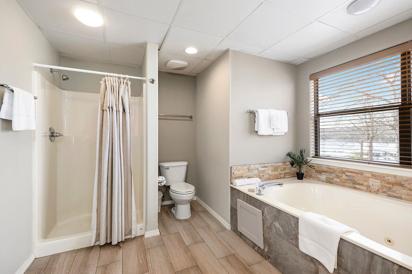 A modern bathroom with a walk-in shower, a toilet, a bathtub, and a window with blinds.