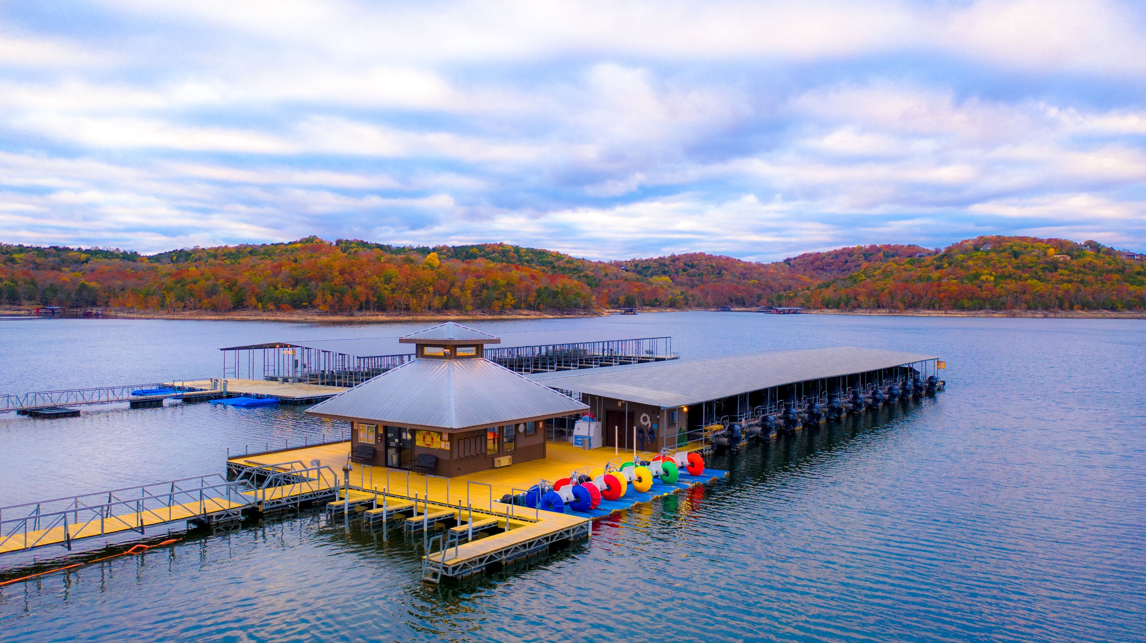 A lakeside dock with a covered building, a floating platform with seating, and a forested hill in the background.