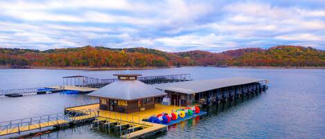 A lakeside dock with a covered building, a floating platform with seating, and a forested hill in the background.