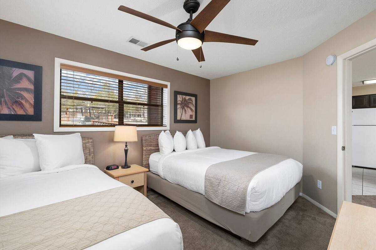 A hotel room with two beds, a ceiling fan, a window with blinds, and a door leading to a kitchen.
