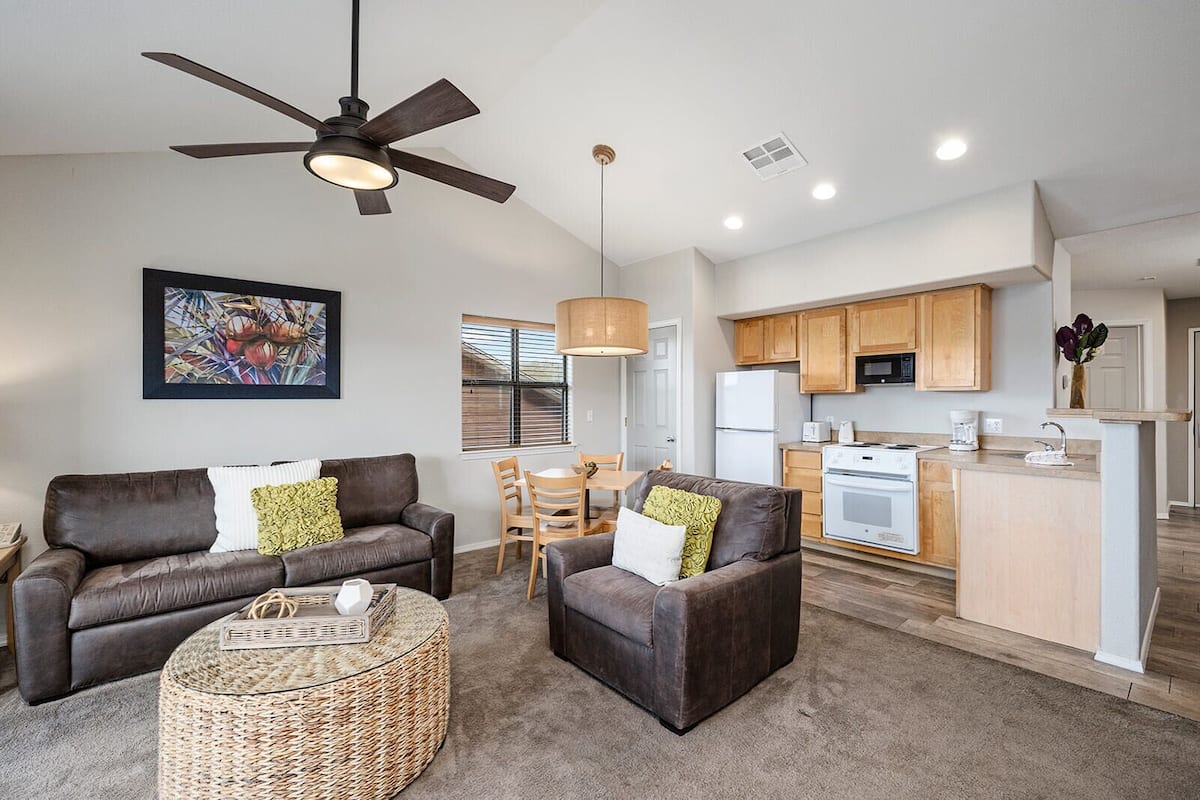 A modern living room with a sofa, armchair, and coffee table, a dining area with a table and chairs, and a kitchen with wooden cabinets and appliances.