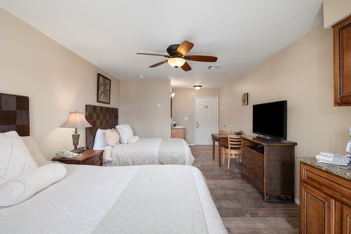 A hotel room with two beds, a desk with a television, a ceiling fan, and a bathroom visible through an open door.