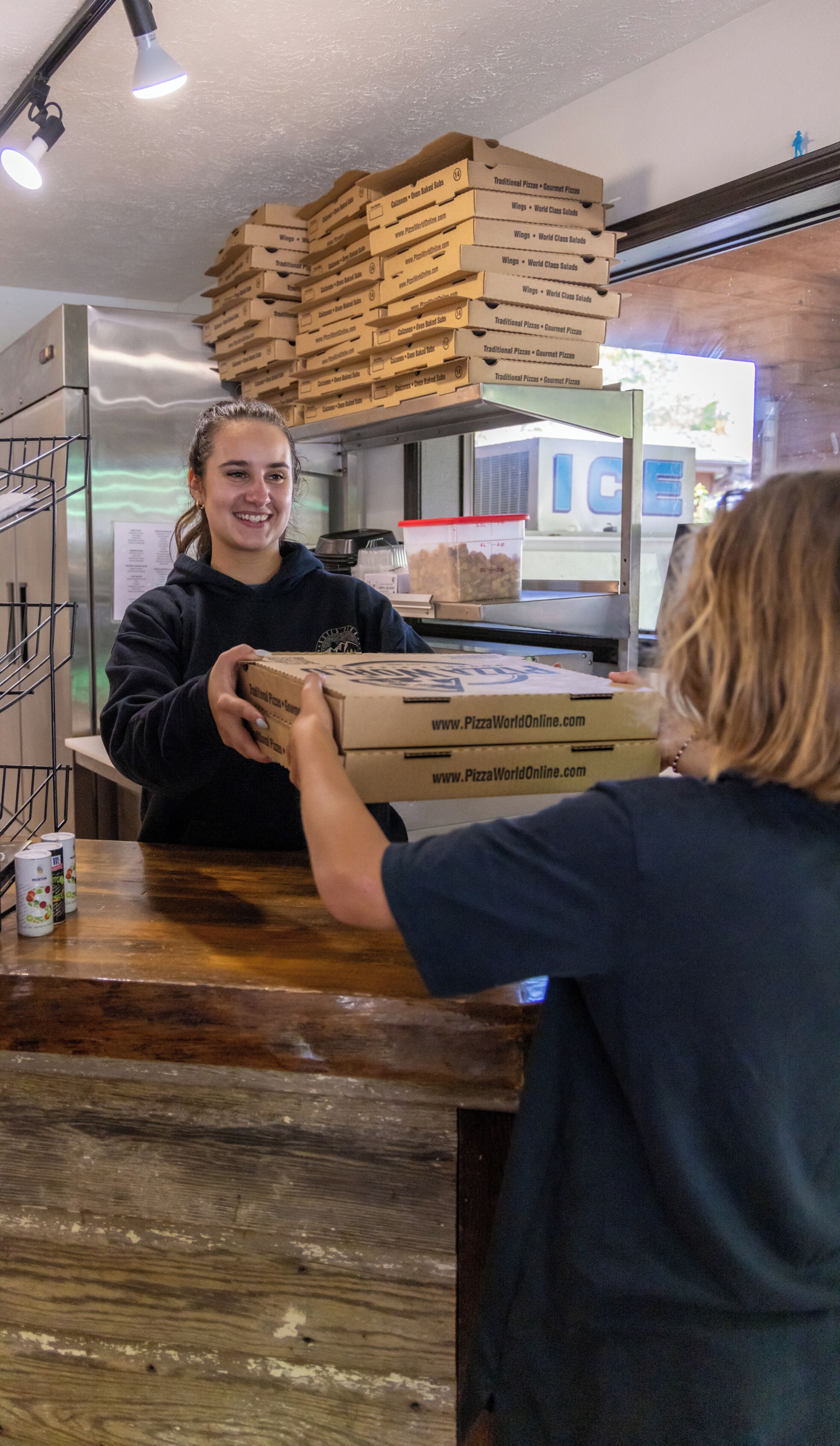 Een persoon geeft een pizzadoos aan een ander persoon bij een balie, met gestapelde pizzadozen op de achtergrond.