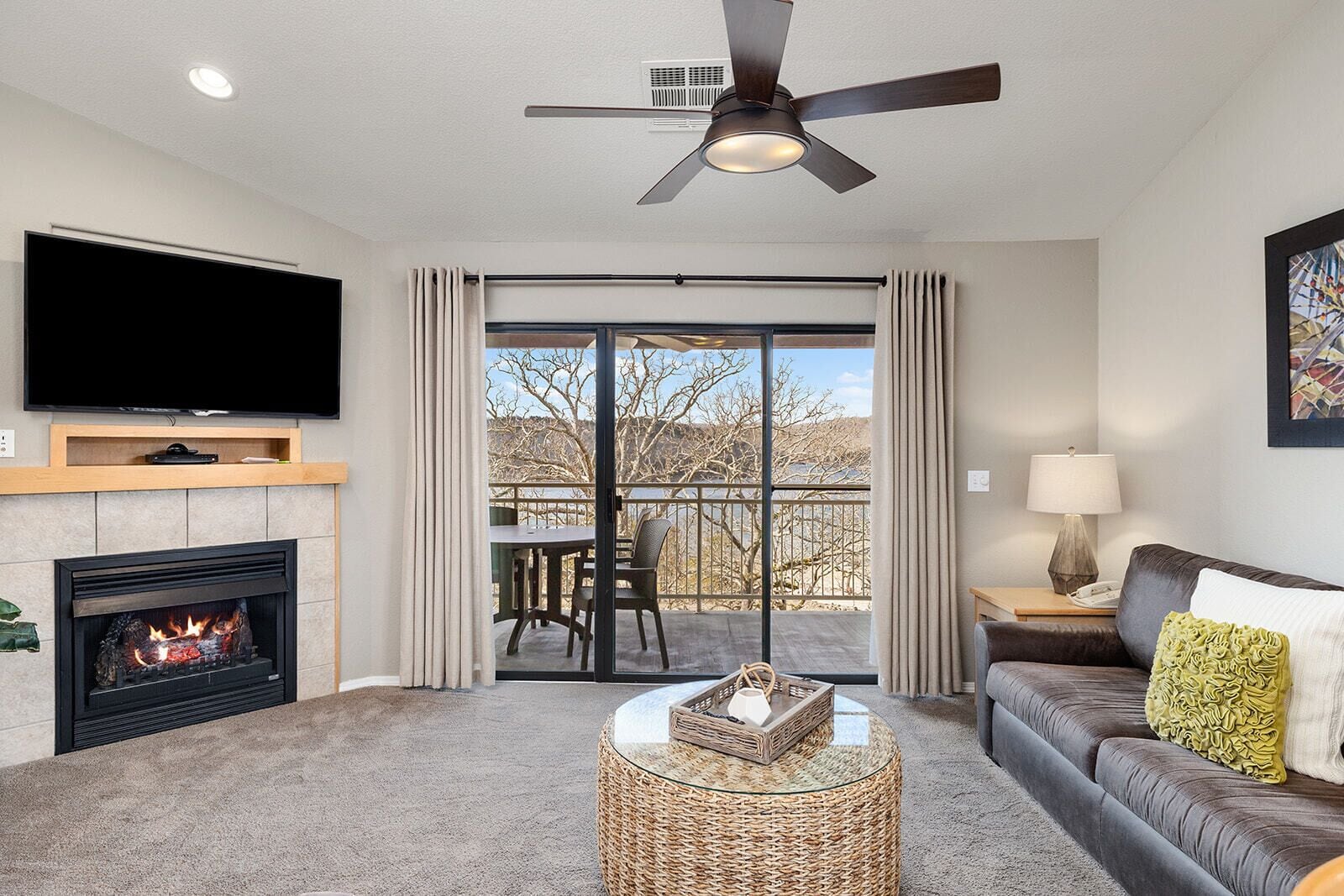 A living room with a fireplace, a flat-screen TV, a ceiling fan, a balcony with a table and chairs, and a sofa with cushions.