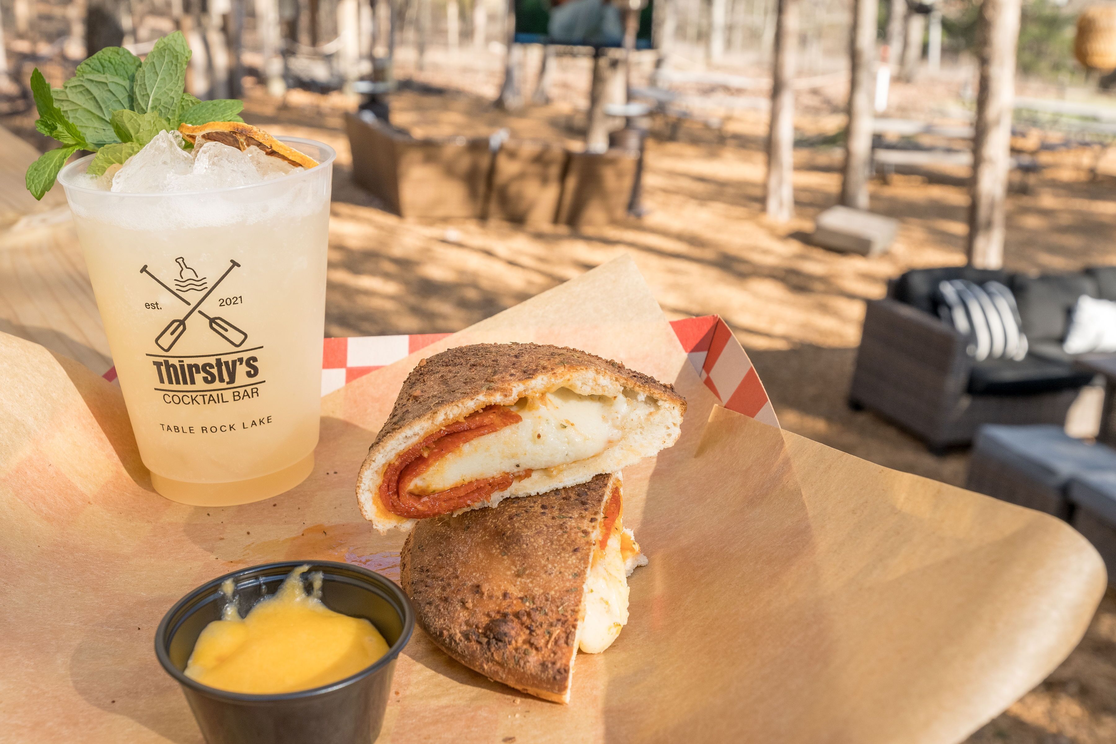 A sandwich with a drink and a small container of butter on a wooden table in an outdoor setting.