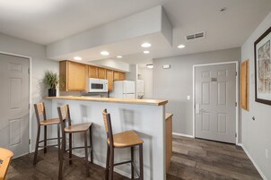 A kitchen with wooden cabinets, a microwave, a refrigerator, and a bar area with three stools.
