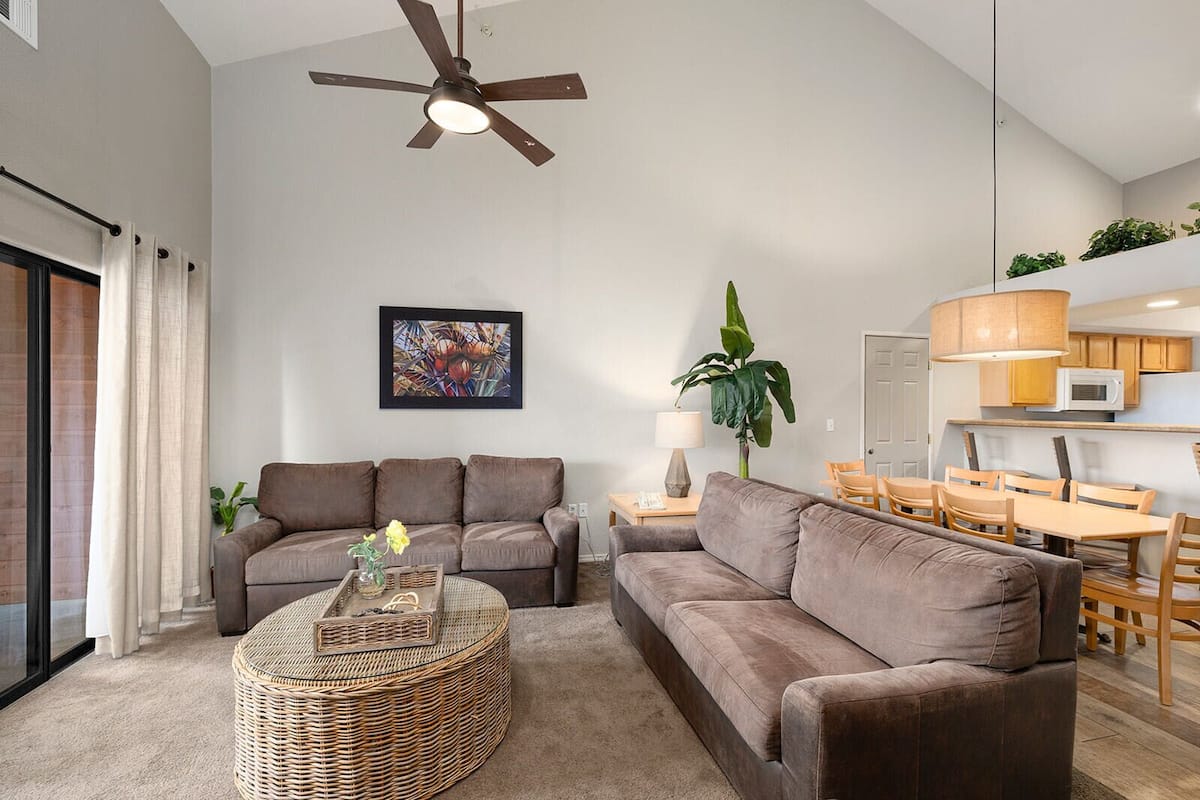 A modern living room with a ceiling fan, a dining area with wooden furniture, and a kitchen with white cabinets.
