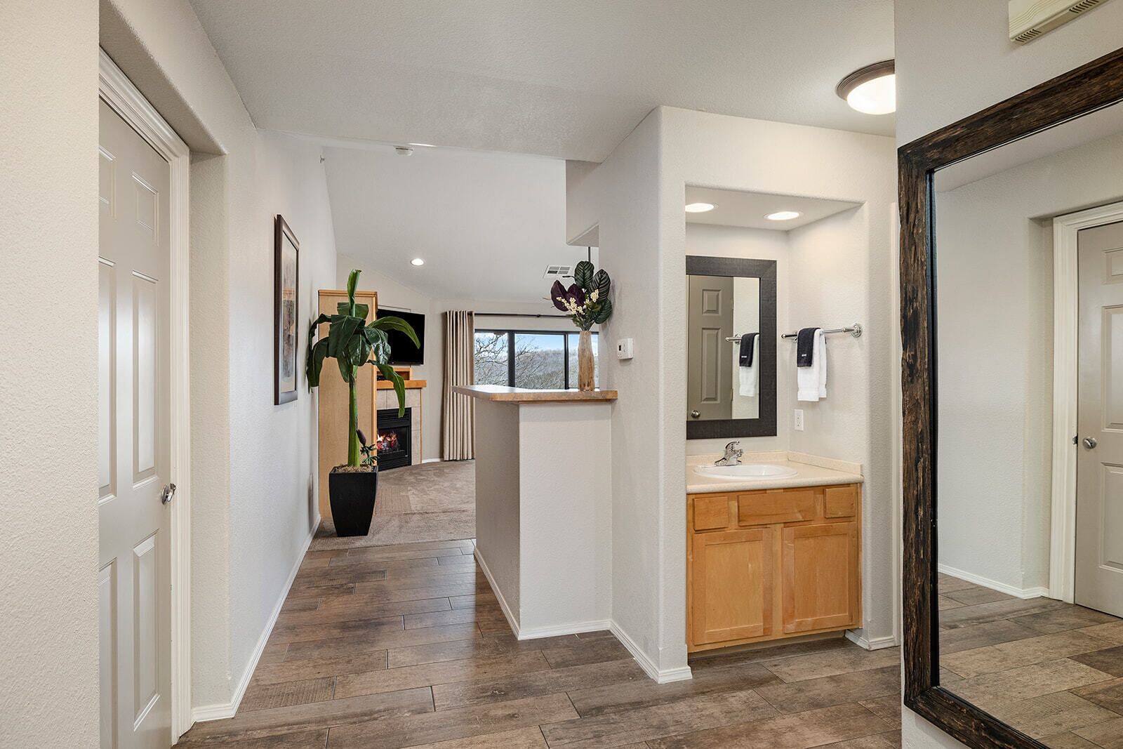 A hallway with wooden flooring, a plant, a fireplace, and a bathroom with a sink and mirror.
