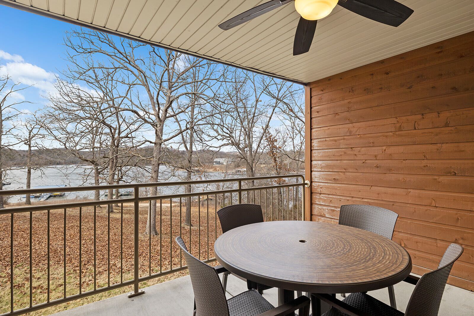 A covered outdoor area with a round table and chairs, a ceiling fan, and a view of trees and a body of water.