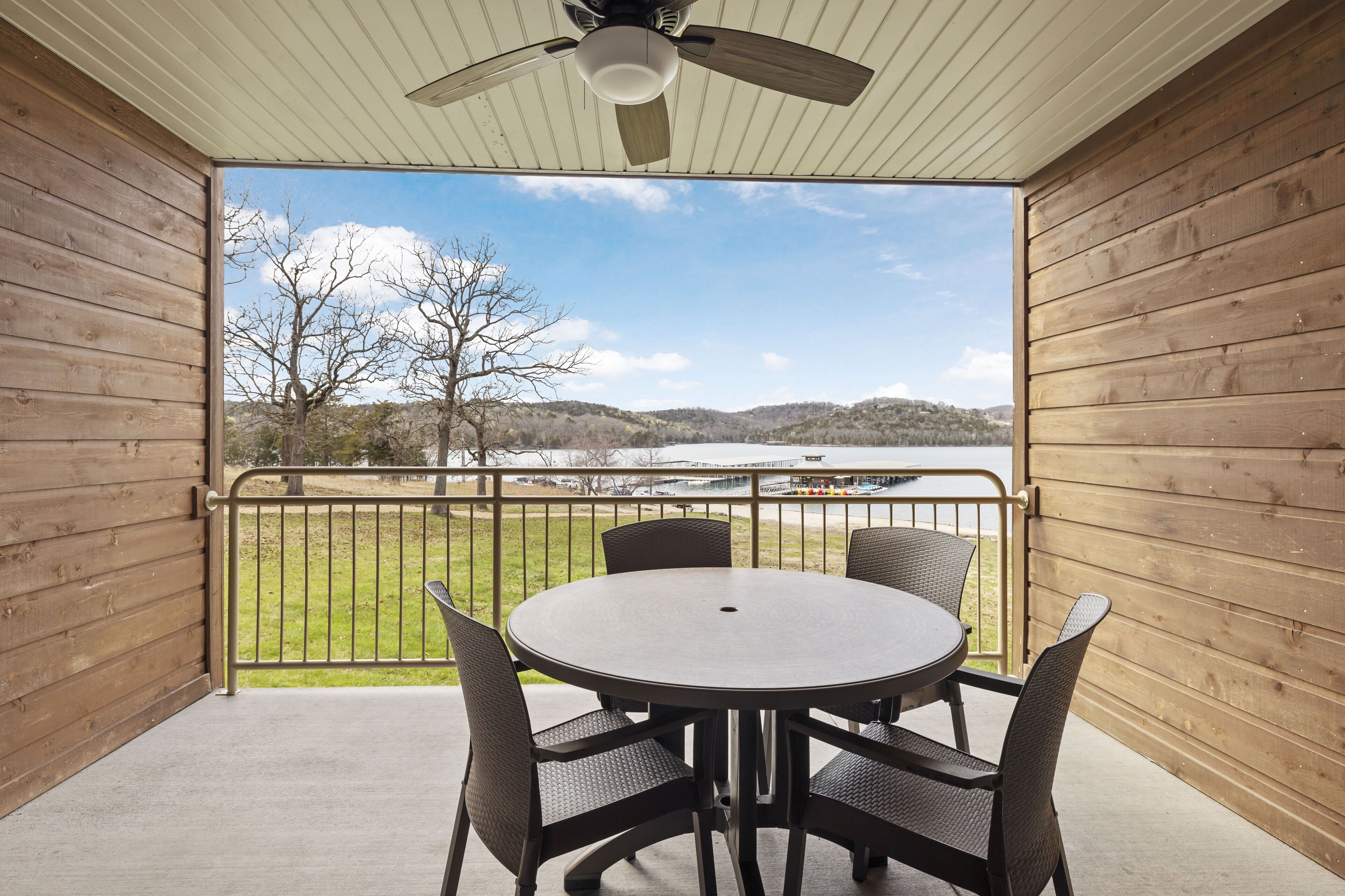 Une terrasse couverte avec une table ronde et des chaises, un ventilateur de plafond et une vue sur un lac et des collines.