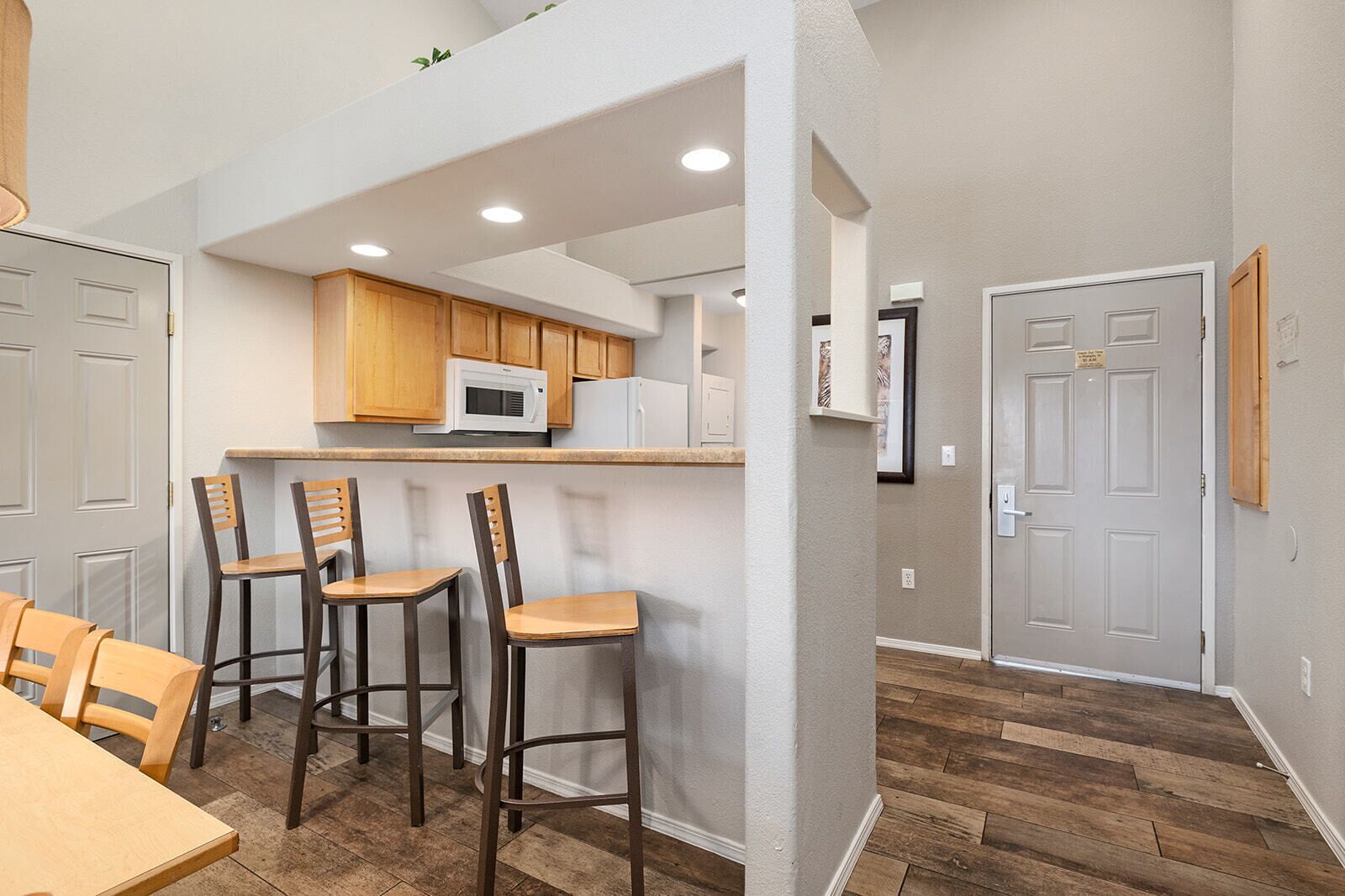 A compact kitchen with wooden cabinets, a microwave, and a refrigerator. There are three bar stools and a dining table with chairs. The floor is wooden, and the walls are painted white.