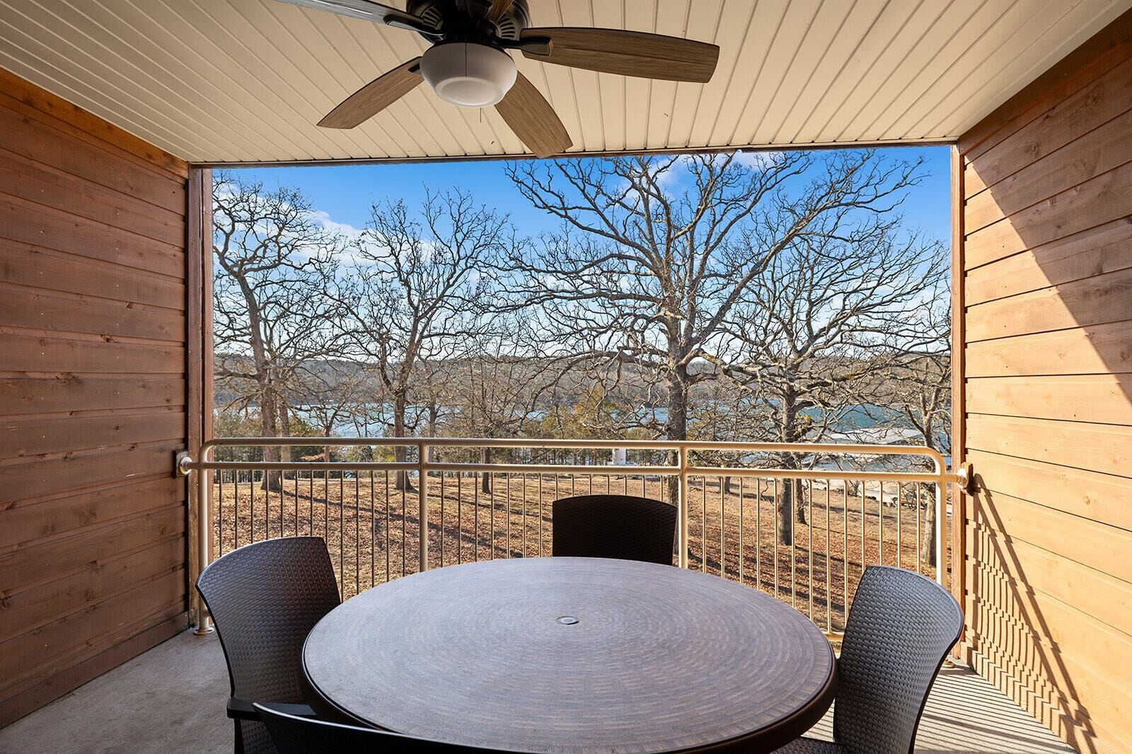 A wooden balcony with a round table and two chairs, overlooking a landscape with trees and a body of water.