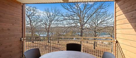 A wooden balcony with a round table and two chairs, overlooking a landscape with trees and a body of water.