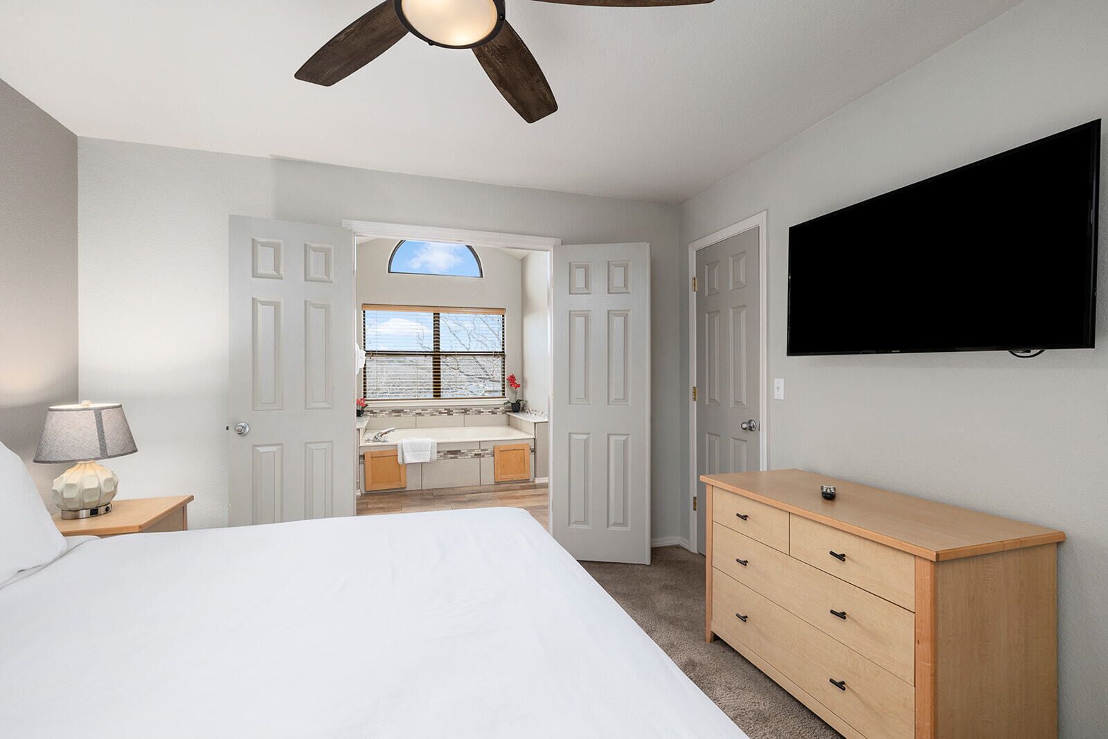 A bedroom with a bed, a nightstand, a dresser, a ceiling fan, and a TV mounted on the wall.