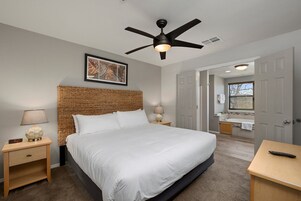 A bedroom with a large bed, two bedside tables, a ceiling fan, a nightstand with a lamp, a framed picture on the wall, and a bathroom visible through an open door.