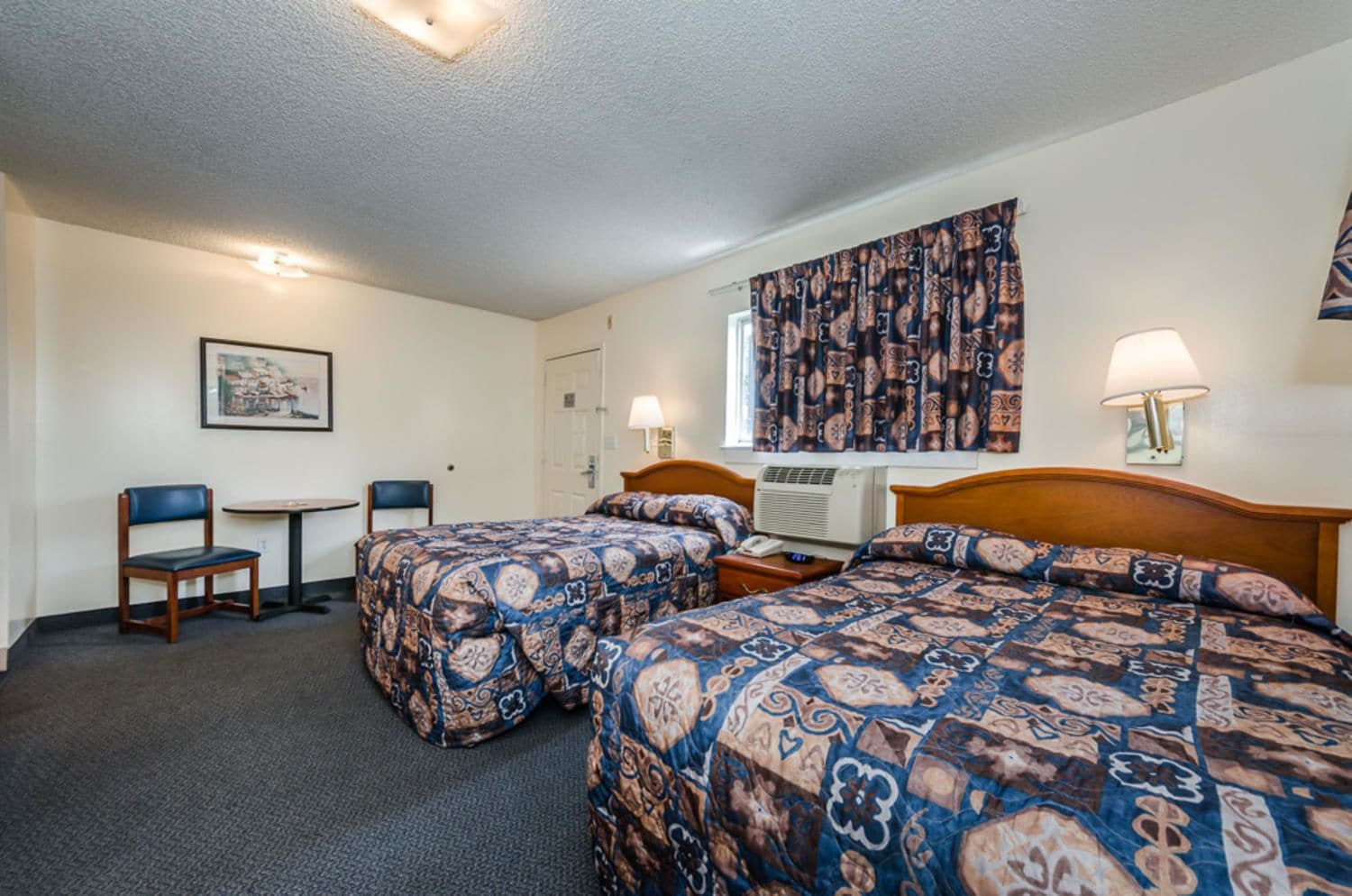 A hotel room with two beds, a desk, a chair, and a window with patterned curtains.