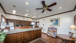 A reception area with a wooden counter, a ceiling fan, a clock, a framed painting, a magazine rack, and a table with chairs.