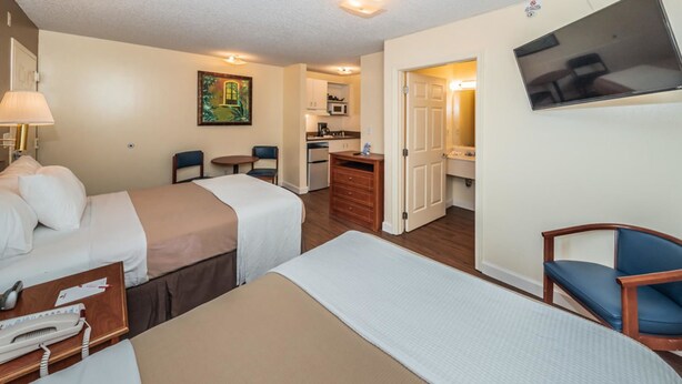 A hotel room with two beds, a desk with a lamp, a chair, a small table, a painting on the wall, a kitchenette, and a bathroom visible through an open door.