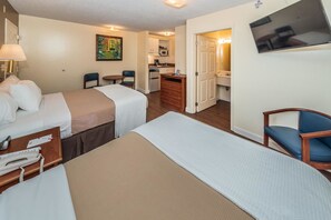 A hotel room with two beds, a desk with a lamp, a chair, a small table, a painting on the wall, a kitchenette, and a bathroom visible through an open door.
