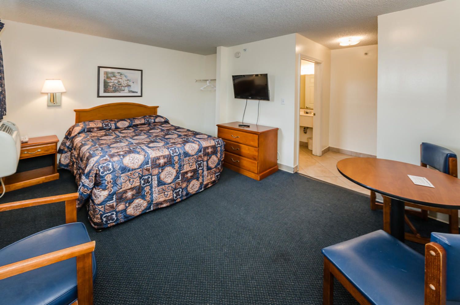 A hotel room with a bed, a desk, a chair, a dresser, a TV, and a small dining table.