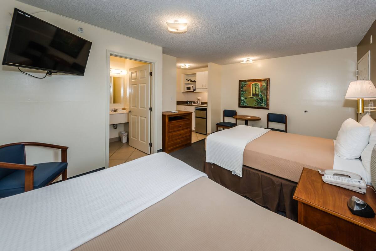 Une chambre d’hôtel avec deux lits, une télévision, un bureau avec un téléphone, une chaise, une petite table et, au fond, une kitchenette.