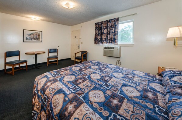 A hotel room with a large bed, two chairs, a small table, an air conditioning unit, and a window with patterned curtains.